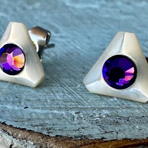 Puede incluir: Un par de pendientes de plata con una base triangular blanca y una piedra preciosa púrpura en el centro.