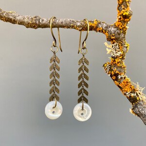 May include: A pair of long, brass earrings with a chevron pattern and a white pearl button at the bottom.