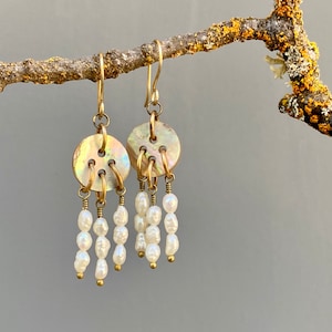 May include: A pair of gold earrings with a round, iridescent button-like charm and a dangling strand of white pearls.
