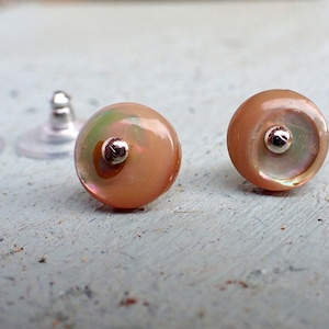 Puede incluir: Un par de pequeños pendientes redondos con un centro de concha iridiscente rosa y un poste plateado. Los pendientes están sobre una superficie azul claro.