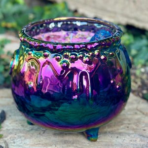 May include: A round, three-legged cauldron with an iridescent, metallic finish that shifts between shades of purple, blue, and green. The exterior features a textured, dripping liquid design around the rim, with a visible blue crystal inside.