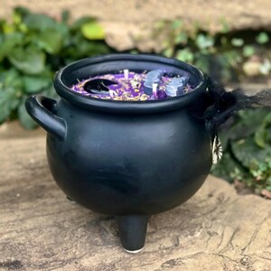 May include: A matte black ceramic cauldron candle with three legs and two side handles. The wax is purple and topped with dried herbs, a large black stone, and grey bat-shaped wax embeds. A black tulle ribbon with a silver charm is tied to one handle.