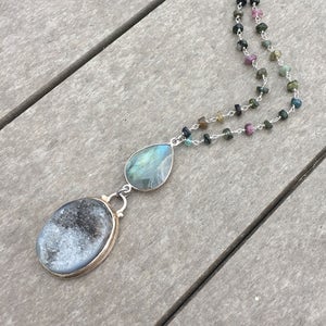 May include: A silver chain necklace with a teardrop-shaped labradorite gemstone and a round, gray agate pendant. The necklace is made with small, multi-colored gemstones.