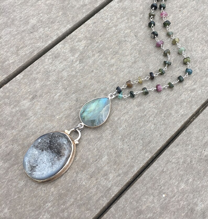 May include: A silver chain necklace with a teardrop-shaped labradorite gemstone and a round, gray agate pendant. The necklace is made with small, multi-colored gemstones.