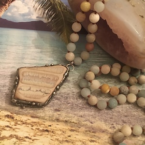 May include: A light blue, brown, and white beaded necklace with a light brown agate pendant. The necklace is draped over a light brown agate stone. The background is a beach scene with a palm tree and blue water.