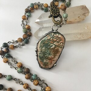 May include: A long necklace with a unique pendant made from a natural stone with a brown, green, and white pattern. The necklace is made with brown, yellow, and turquoise beads and a silver chain.