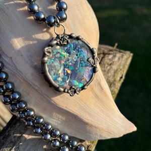 May include: A silver necklace with a round iridescent glass pendant. The pendant is set in a silver frame with decorative details. The necklace is made of black beads.