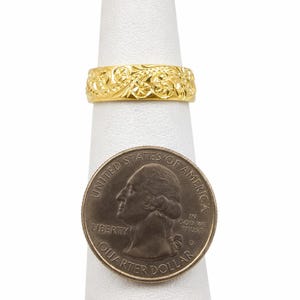 May include: A gold-colored ring with an intricate floral design. The ring is displayed on a white ring holder next to a U.S. quarter coin. The ring's detailed pattern is visible, showcasing its craftsmanship. The quarter coin is included for size reference.