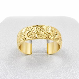 May include: A gold-colored ring with an intricate floral and scrollwork design. The ring has a wide band with a detailed pattern that wraps around the entire circumference. The ring is displayed on a white surface.