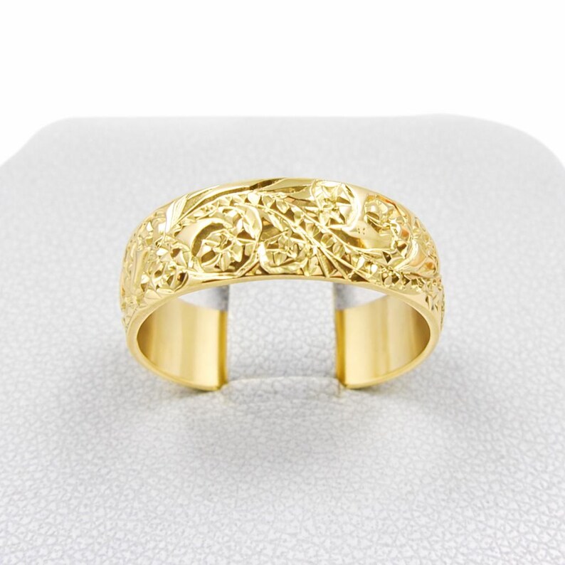 May include: A gold-colored ring with an intricate floral and scrollwork design. The ring has a wide band with a detailed pattern that wraps around the entire circumference. The ring is displayed on a white surface.