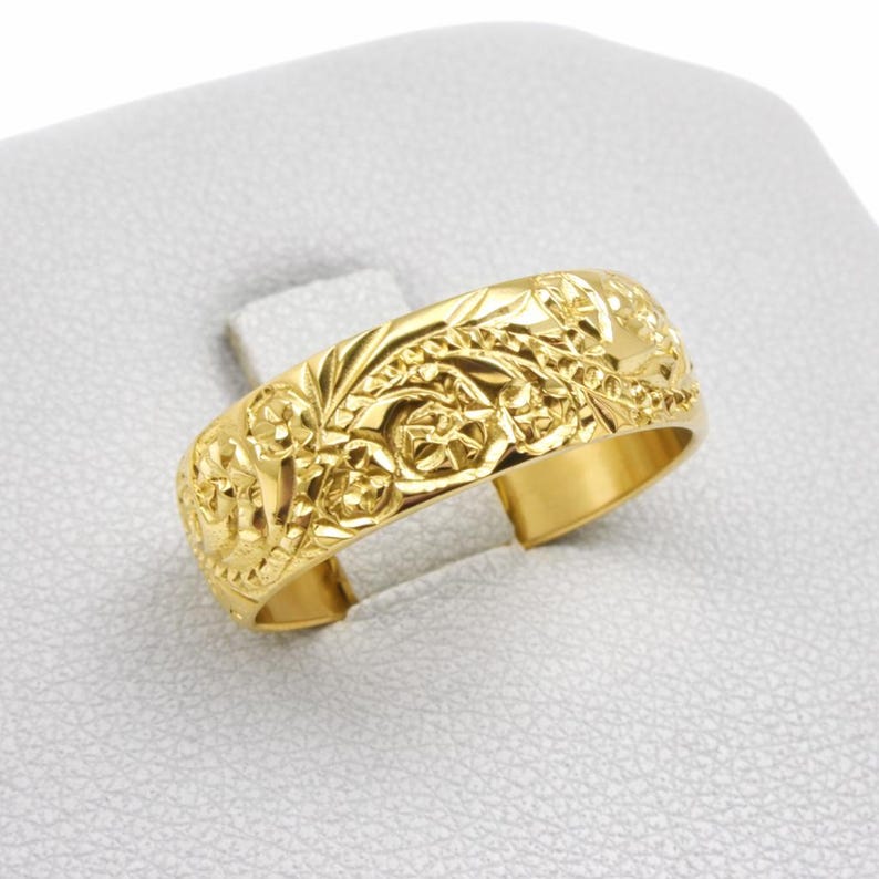 May include: A gold-toned ring featuring an intricate, embossed floral pattern across its entire outer surface. The band has a polished, reflective finish with detailed engravings of leaves and blossoms, creating a textured appearance.