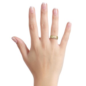 May include: A gold-colored ring with an intricate, textured design is worn on a hand. The ring appears to be a band style, with the detailed pattern covering the entire surface. The ring is likely made of metal and is displayed against a white background.