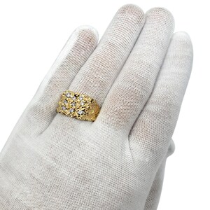 May include: A gold ring featuring a textured, nugget-style surface with a rectangular top section. The ring is set with nine small, clear, round-cut stones arranged in a three-by-three grid pattern across the center of the textured gold band.