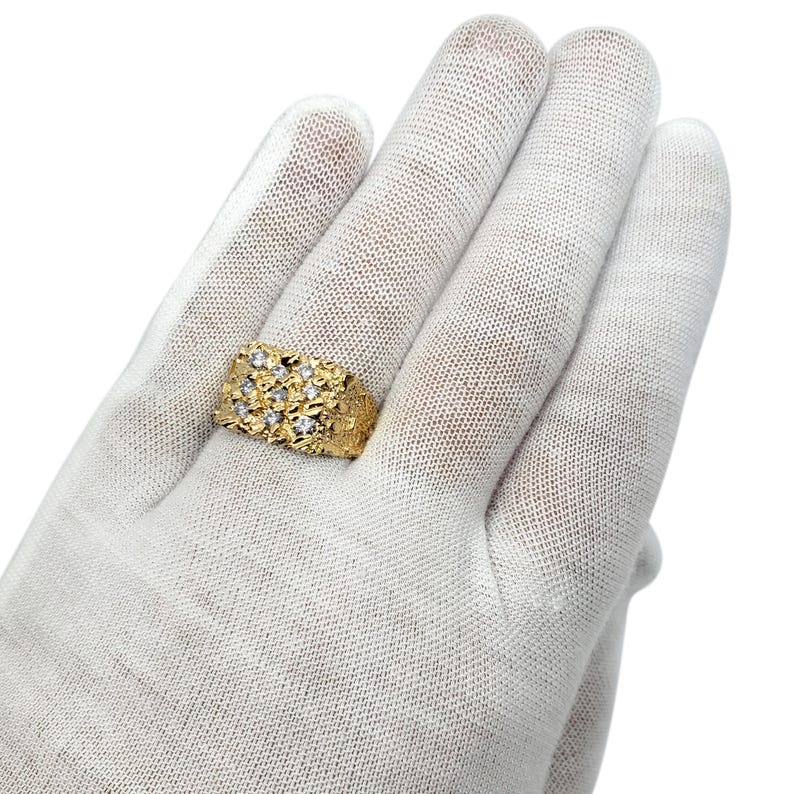 May include: A gold ring featuring a textured, nugget-style surface with a rectangular top section. The ring is set with nine small, clear, round-cut stones arranged in a three-by-three grid pattern across the center of the textured gold band.