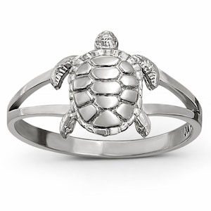 May include: A polished silver-toned ring featuring a central sea turtle motif. The turtle has a detailed, segmented shell and extended flippers that connect to a split-shank band. The metal surface is smooth and reflective throughout the design.