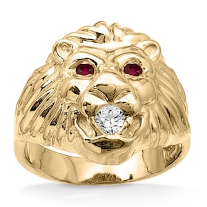 May include: A gold-colored lion head ring with ruby eyes and a diamond in its mouth. The ring is a detailed sculpture of a lion's face, showcasing its mane and facial features. The ring is set against a white background.
