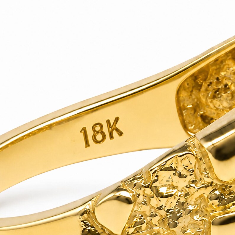 May include: A close-up view of the interior band of a gold ring, featuring the stamped text 18K. The metal surface is smooth and polished, contrasting with the textured, nugget-style finish visible on the outer edge of the ring band.