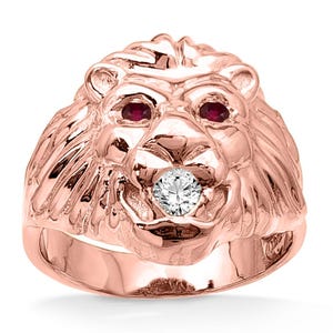 May include: A rose gold lion head ring with ruby eyes and a diamond in its mouth. The ring is a detailed sculpture of a lion's face, showcasing the mane and facial features. The ring is a statement piece of jewelry.