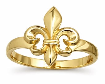 May include: A polished yellow gold ring featuring a prominent fleur-de-lis emblem centered on the band. The metal surface is smooth and reflective, showcasing the classic three-petaled design with a central vertical element and two curved side petals.