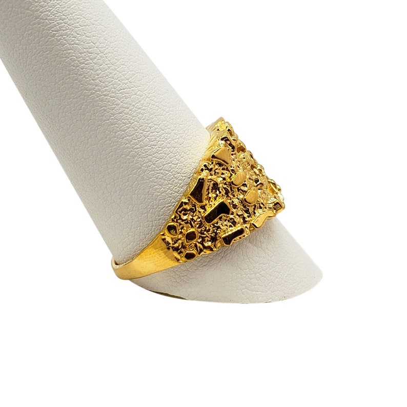 May include: A gold ring with a textured, nugget-like design. The ring is displayed on a white ring stand.