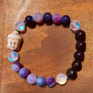 Puede incluir: Pulsera de cuentas con una cabeza de Buda blanca, cuentas iridiscentes y varias cuentas de piedra moradas y negras. La pulsera está ensartada en un cordón elástico plateado y se muestra sobre una superficie dorada.