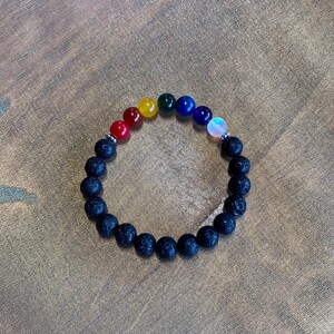 Puede incluir: Una pulsera de cuentas con una combinación de cuentas de roca de lava negra y piedras de chakra de colores. Las piedras de chakra están dispuestas en un espectro de arcoíris, con colores rojo, naranja, amarillo, verde, azul, índigo y violeta. La pulsera está sobre una superficie dorada.