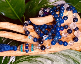 May include: A beaded necklace with deep blue lapis lazuli and clear quartz beads. The necklace has a decorative silver pendant with turquoise and coral accents, and a blue tassel. The beads are displayed on a wooden hand.