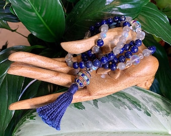 May include: A mala necklace with a navy blue tassel. The necklace features dark blue lapis lazuli beads, grey labradorite beads, and clear quartz beads. The tassel is navy blue and the decorative bead is multi-coloured.
