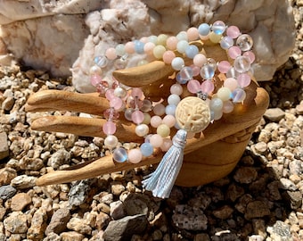 May include: A beaded mala necklace with a carved ivory pendant and a light blue tassel. The necklace features a mix of pink, white, and blue beads, including pearls and crystal accents. The beads are strung together, creating a layered effect.