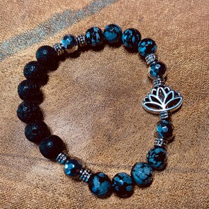 May include: A beaded bracelet featuring black lava rock beads, snowflake obsidian beads, and faceted dark gray beads. The bracelet includes silver-toned spacers and a silver lotus flower charm. The bracelet is designed for wrist sizes up to 7.5 inches.