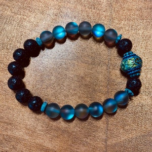 Peut inclure: Un bracelet de perles composé de perles de roche volcanique noire, de perles de verre bleu-vert givré et d'une perle décorative bleue et dorée. Le bracelet est monté sur un cordon élastique et est présenté sur une surface dorée texturée.