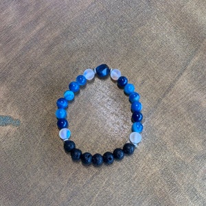 Könnte beinhalten: Ein Armband aus blauen, weißen und schwarzen Perlen. Das Armband hat verschiedene Blautöne, mit weißen und schwarzen Lavaperlen. Die Perlen sind auf einem Gummiband aufgefädelt. Das Armband hat einen Umfang von etwa 17 cm.