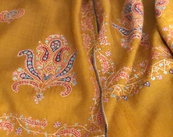May include: A mustard yellow shawl with intricate paisley embroidery in shades of pink, blue, and white. The shawl has a fringed border.