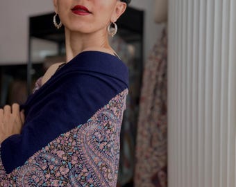 May include: A navy blue shawl featuring an intricate, multi-colored paisley pattern in shades of pink, light blue, and cream. The garment is draped over the shoulders, showcasing a solid navy blue section at the top and a detailed, ornate floral-inspired paisley design covering the lower portion of the fabric.