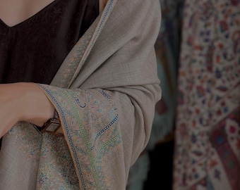 May include: A light beige shawl featuring an intricate embroidered border with blue, green, and orange floral patterns. The shawl is draped over a black camisole, showcasing the detailed needlework along the edges and the soft texture of the fabric.