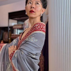 May include: A grey shawl featuring a wide, deep red border adorned with intricate white and light blue floral embroidery. The fabric displays a smooth texture, and the edges are finished with a subtle, thin black and white striped pattern.