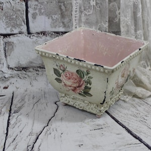 May include: Square, cream-coloured planter with a distressed finish, featuring a pink interior and rose floral decals. The planter has a decorative base and is set against a rustic, white-washed brick wall and wooden surface.