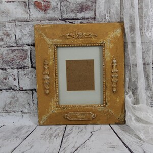 May include: A gold-painted picture frame with ornate details and a white mount. The frame is sitting on a whitewashed wood surface.