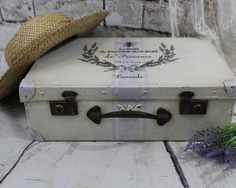 May include: A white vintage suitcase with a lavender and white striped design. The suitcase has a metal handle and two locks. The text "Les Aromates de Provence Miel & Lavande" is printed on the top of the suitcase. The suitcase is sitting on a white wooden surface.