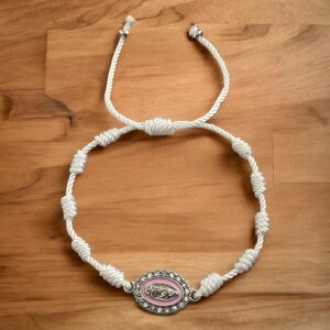 May include: White knotted cord bracelet with a silver oval charm featuring a pink enamel image of the Virgin Mary.