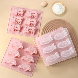 May include: Three pink silicone molds for baking. The molds are shaped like trains, cars, and airplanes.