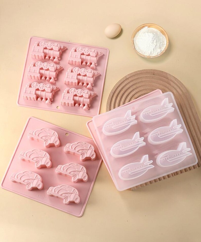 May include: Three pink silicone molds for baking. The molds are shaped like trains, cars, and airplanes.