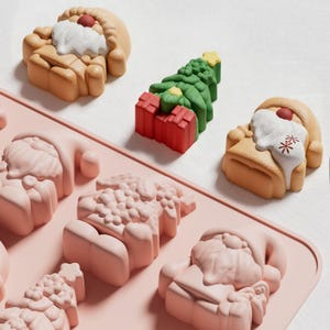 Puede incluir: Un molde de silicona rosa con formas navideñas. El molde presenta figuras de Papá Noel con barbas blancas y narices rojas, y árboles de Navidad con cajas de regalo rojas. Las formas están en varias etapas de finalización, algunas completas.