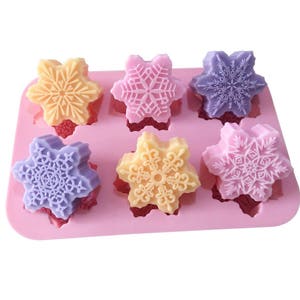 Può includere: Uno stampo in silicone rosa con sei cavità a forma di fiocco di neve. Lo stampo è riempito con sei saponette colorate, ciascuna con un diverso design a fiocco di neve. Le saponette sono in tonalità di viola, giallo e arancio.