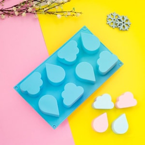 Pode incluir: Um molde de silicone azul claro com várias formas, incluindo gotas de chuva e nuvens. O molde está sobre um fundo rosa e amarelo, com duas decorações de floco de neve e várias peças de silicone em forma de nuvem e gota de chuva.
