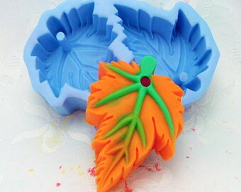 Puede incluir: Un molde de silicona azul con forma de dos hojas, con una hoja naranja y verde hecha de resina sentada encima del molde.