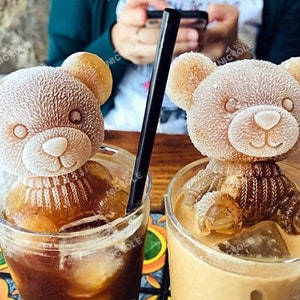 May include: Two iced coffee drinks with frozen teddy bear ice cubes. The teddy bears are wearing brown sweaters and are sitting in the drinks. The drinks are in clear glasses with straws.
