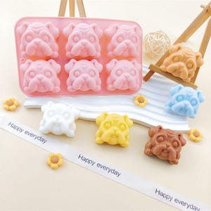 May include: A pink silicone mold with six pug-shaped cavities. Several pug-shaped soaps in various colors, including white, yellow, blue, brown, and orange, are displayed. The phrase "Happy everyday" is printed on a white ribbon.