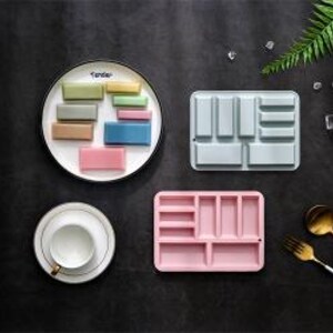 May include: A white plate with the word Tender printed on it, holding various rectangular blocks in shades of green, blue, pink, and yellow. Beside it are a light blue and a pink rectangular silicone mould, a white teacup, and gold cutlery.