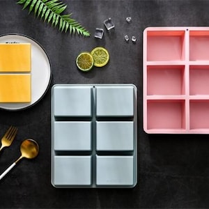 May include: Two rectangular silicone moulds with six square compartments each, one in light blue and one in pink. A white plate holds two yellow square blocks, accompanied by a gold fork and spoon, a green leaf, two lime slices, and four ice cubes.